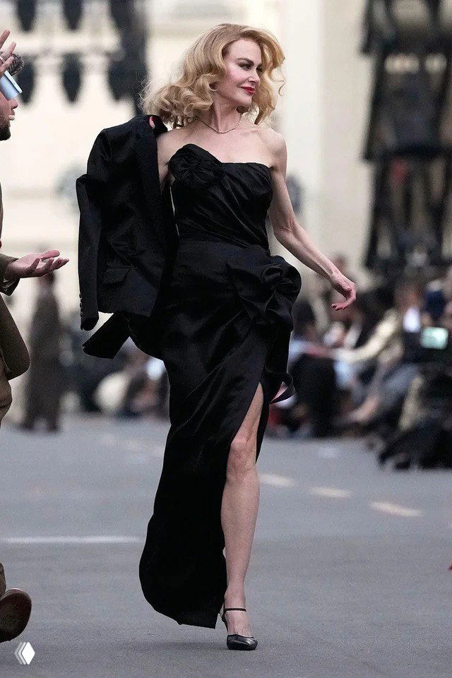 Актриса снимает жакет, демонстрируя чёрную драпировку платья с крупным декоративным узлом и высоким разрезом, вокруг — камеры и зрители.