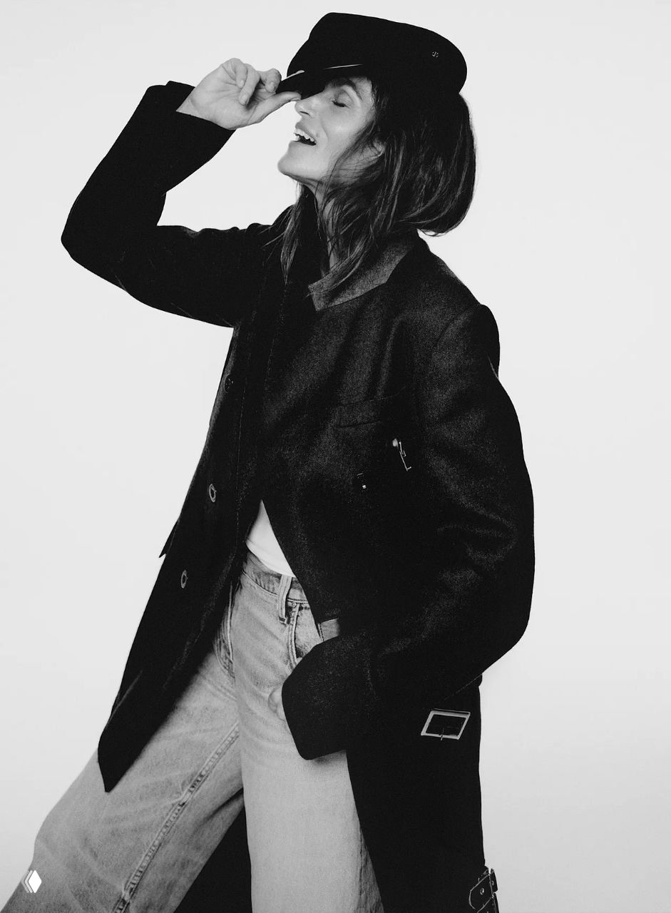 Чёрно‑белый кадр: модель в длинном пальто и шляпе смеётся в студии — динамичная поза и акцент на силуэте одежды.