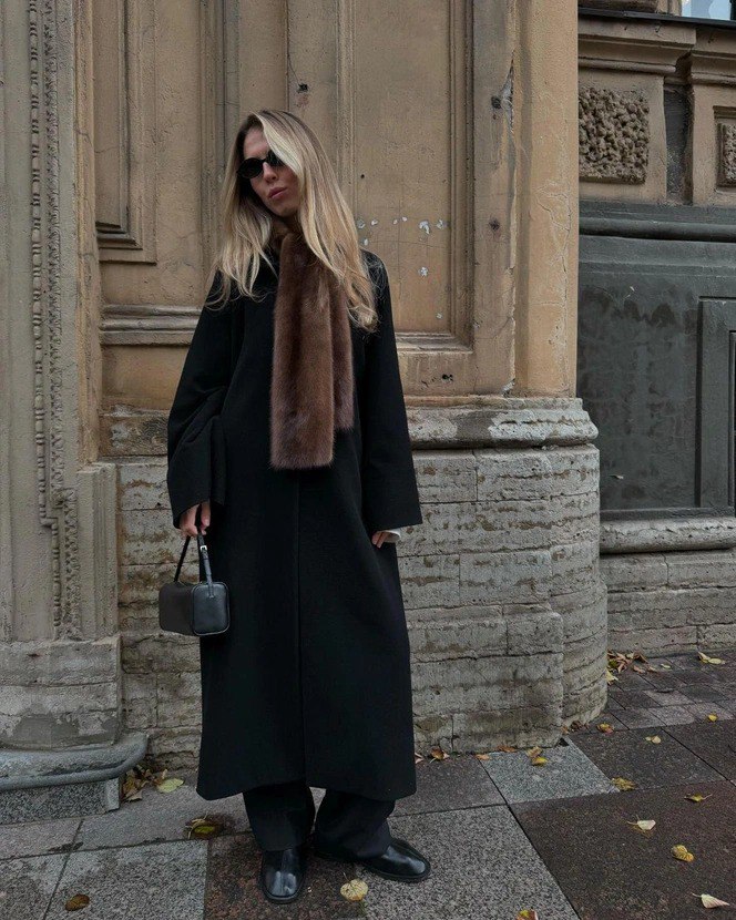 Стритстайл: женщина в длинном чёрном пальто и коричневом меховом шарфе на фоне архитектуры города — пастельная палитра и минимализм.