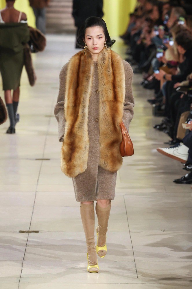 Модель в пальто с широким рыжим меховым горжетом идёт по подиуму; акцент на объемный роскошный аксессуар в образе осень-зима.
