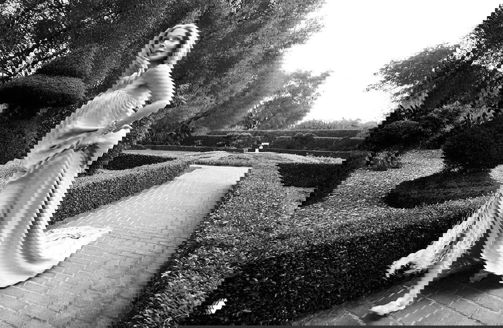 Черно-белая фотография: женщина в длинном платье идёт по вымощенной дорожке в формальном саду для глянцевого издания, съёмка на природе.