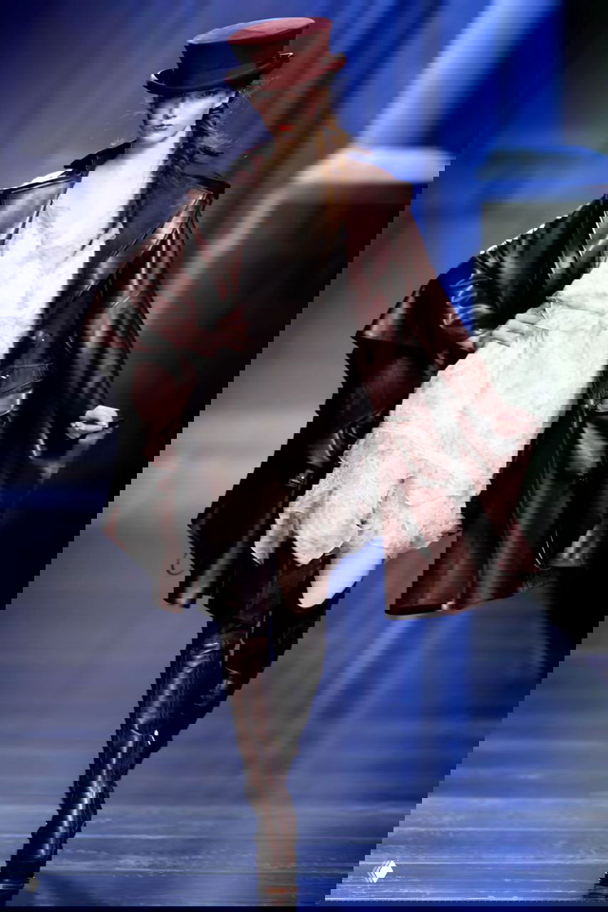 Модель в тёмно‑бордовом пальто из лакированной кожи и мини‑платье, контраст текстуры кожи и лёгких тканей на подиуме Dior AW 2010.