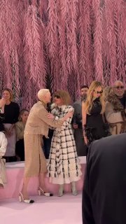 Гости в первом ряду показа Chanel обмениваются объятиями и приветствиями у сцены; видны зрители, дорожка и розоватая инсталляция в оформлении шоу.