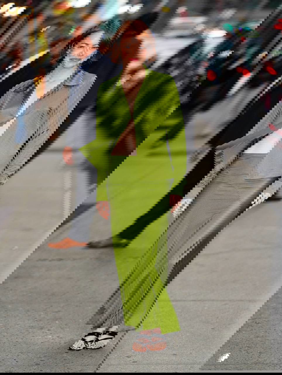 Актриса в оливково‑зелёном шелковом костюме от Calvin Klein идёт по городскому тротуару, улыбается фотографам в вечерней обстановке.