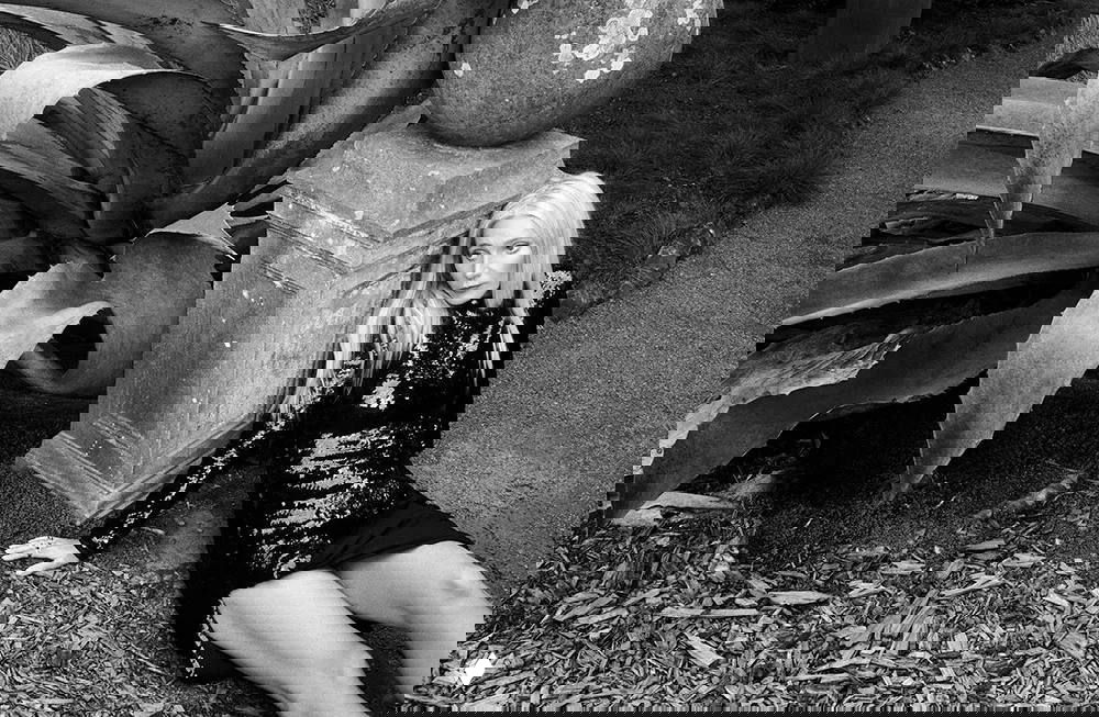 Черно-белое фото: женщина в блестящем платье прислонилась к каменной колонне в садовой локации — строгий модный портрет для журнала.