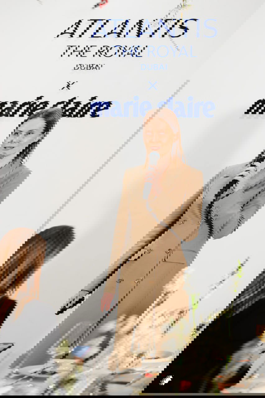 Спикер на сцене рассказывает о философии отеля Atlantis The Royal, женщина с микрофоном выступает на фоне логотипа Atlantis и Marie Claire перед гостями за столами.