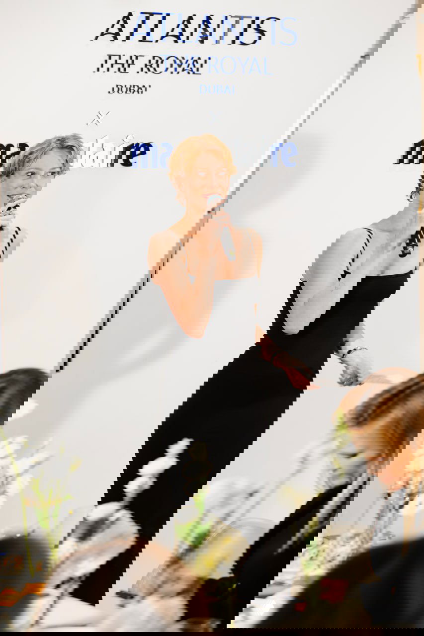 Женщина в чёрном платье держит микрофон и выступает перед гостями за столом, на переднем плане столовая сервировка и цветы, атмосфера делового ужина.