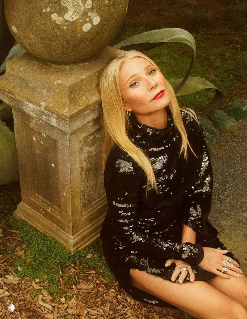 Тёплый портрет: женщина в блестящем тёмном платье сидит у каменной базы с украшениями на руках, модный портрет в садовой локации для журнала.