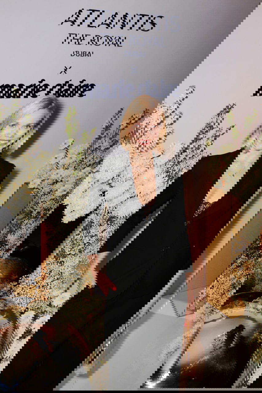 Портретная фотография женщины в тёмном костюме на фоне бренд-зоны Atlantis The Royal x Marie Claire и крупных белых цветочных инсталляций.