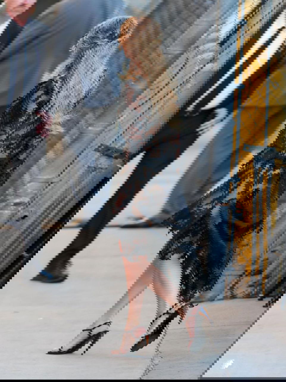 Задний план: Марго Робби в кружевном чёрном платье у ограждения и службы безопасности, видно движение и атмосферу съёмочного района.