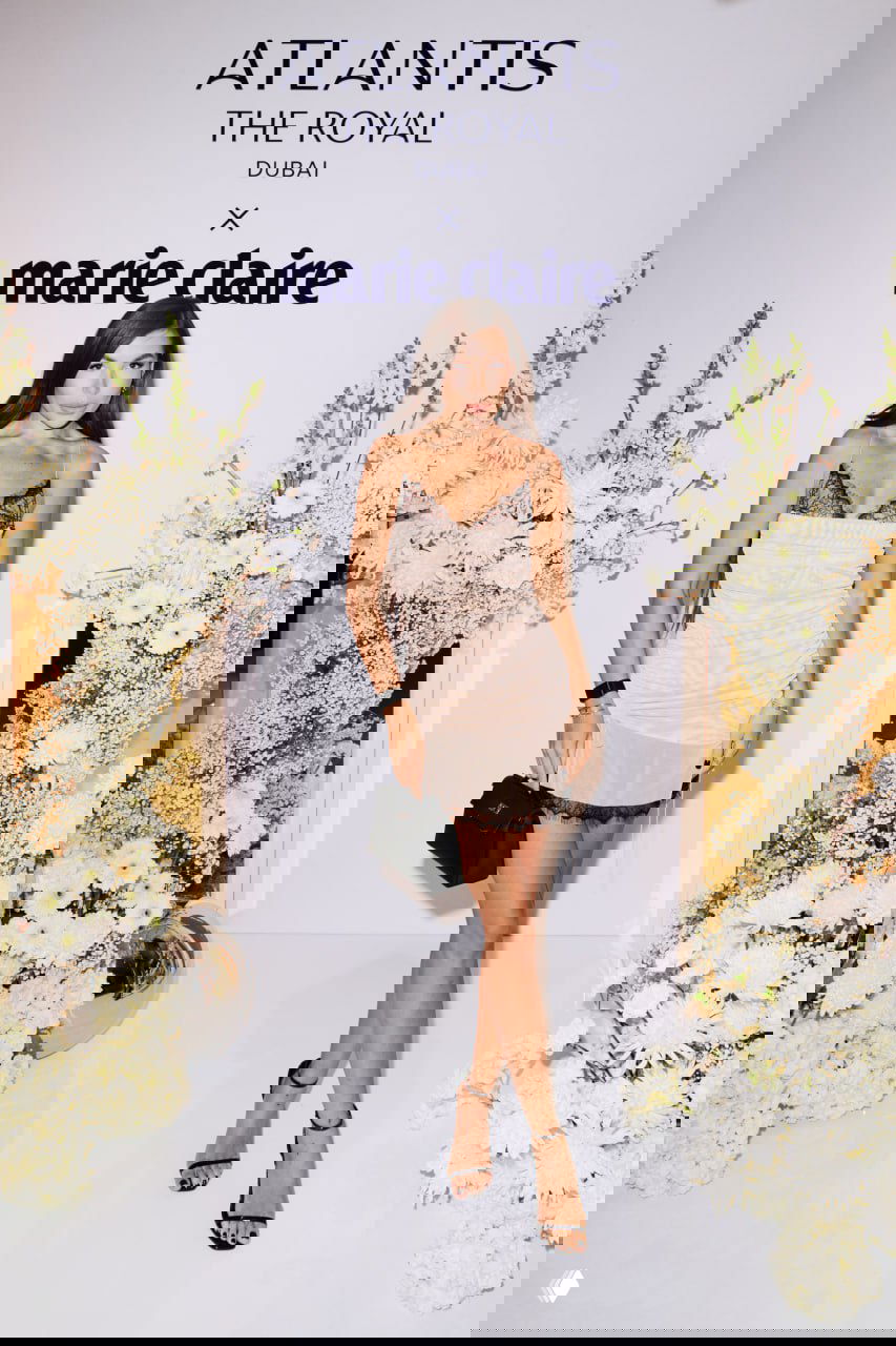 Модель в светлом коктейльном платье позирует у фотозоны Atlantis The Royal x Marie Claire, вокруг белые цветы и сценографические элементы декора.