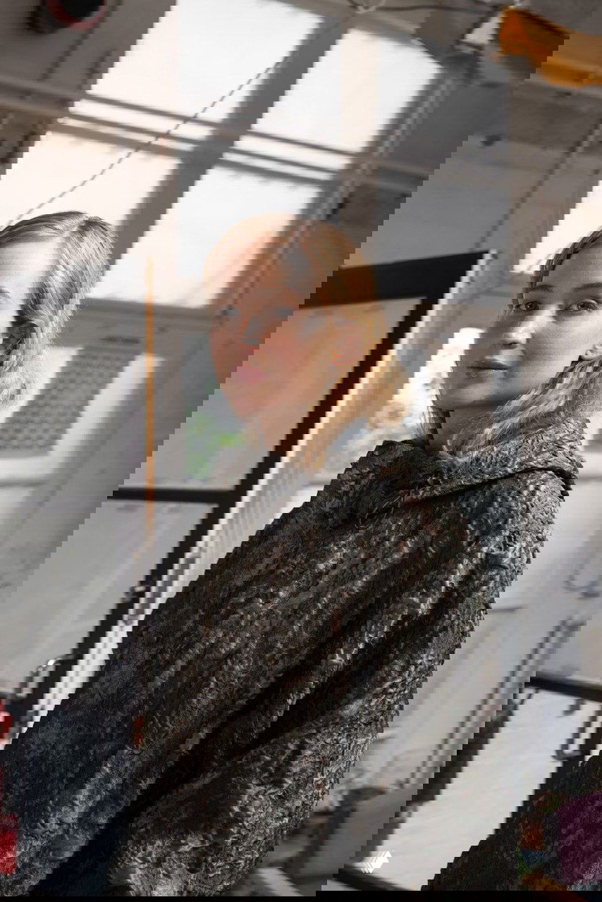 Портрет в индустриальном интерьере: женщина в тёмной фактурной куртке на фоне окна мастерской — текстура и атмосфера пространства.