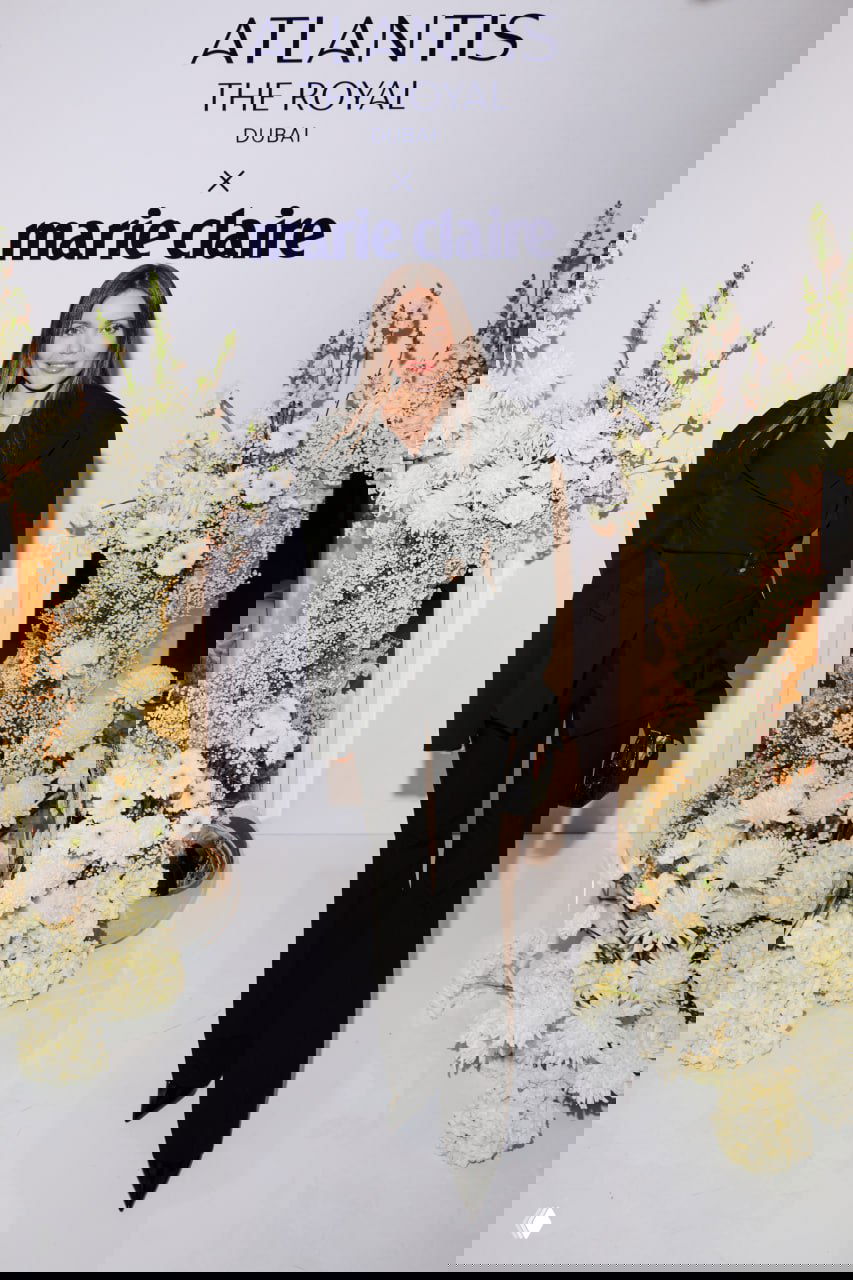 Ещё одна гостья вечера в элегантном костюме позирует перед бренд-бэкдропом Atlantis The Royal x Marie Claire, фотография сделана в студийном стиле с цветами.