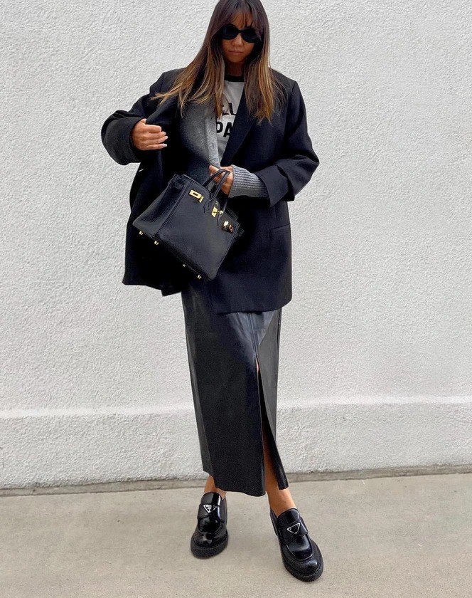 Уличный стритстайл: длинная тёмная юбка‑карандаш из кожи, оверсайз пиджак, чёрная сумка и лаковые мокасины на светлом фоне стены.