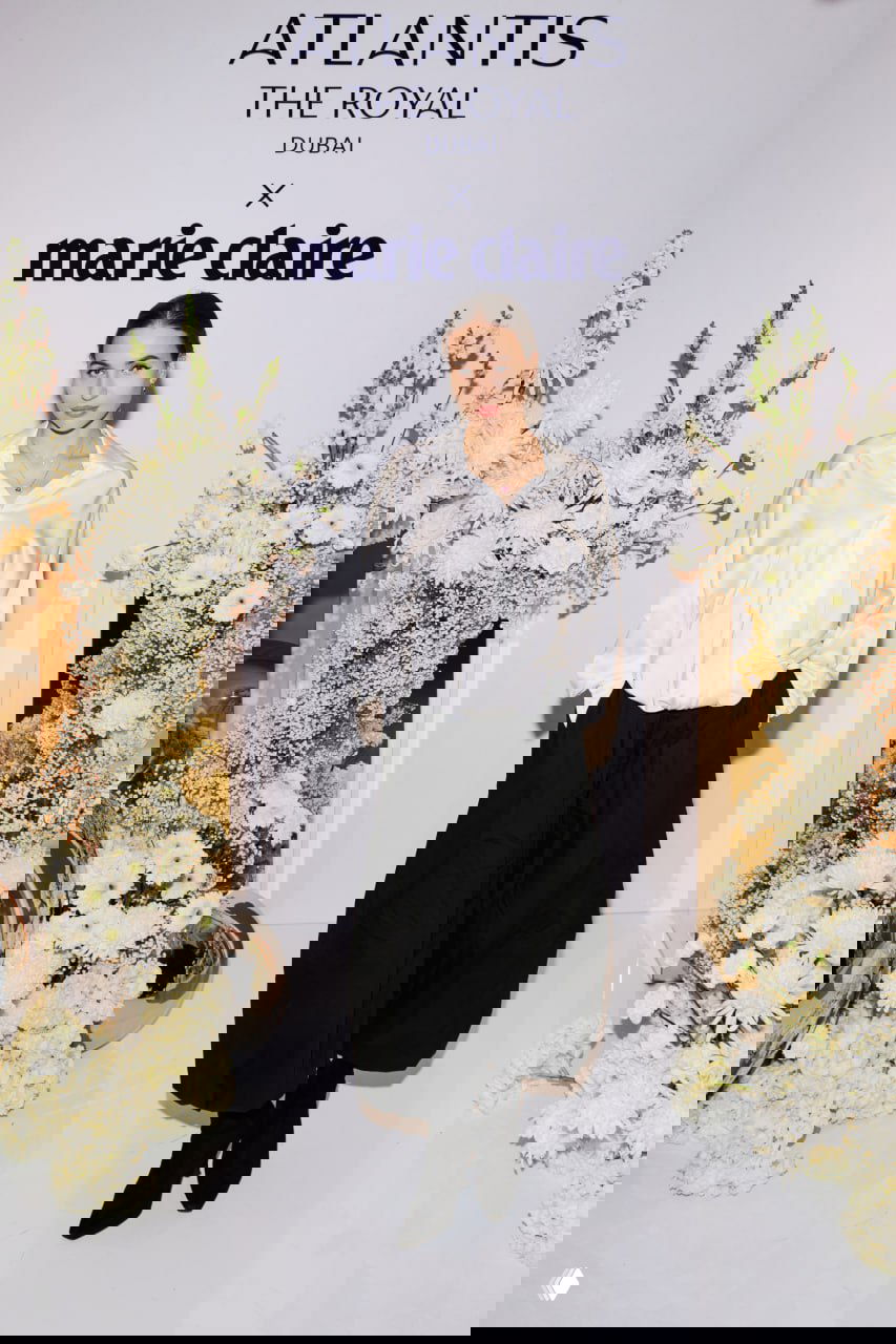 Женщина в чёрном костюме выходит к камере у логотипа Atlantis The Royal x Marie Claire, пол и окружение украшены белыми цветами и декоративными шарами.