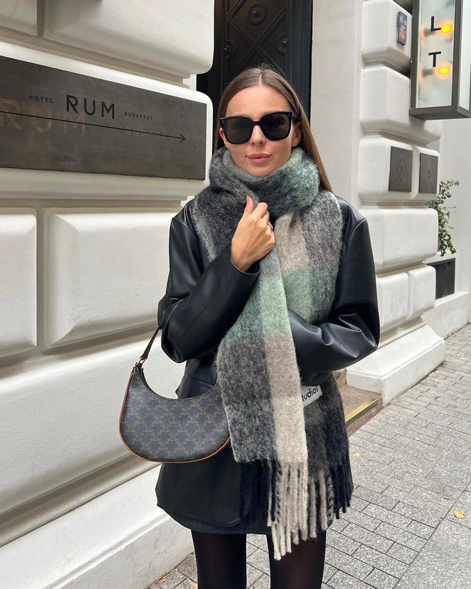 Женщина в кожаной куртке и крупном мягком плед-шарфе в пастельных тонах; городской фон подчёркивает повседневную элегантность образа.