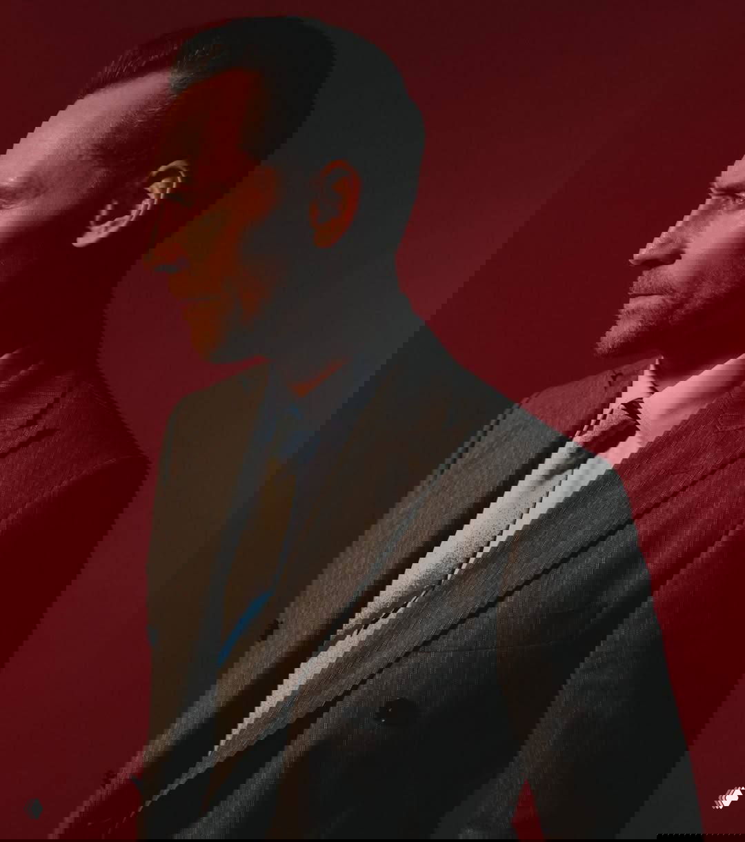 Профильный портрет мужчины в пиджаке на красном фоне, подчёркнутые контуры лица и сдержанная поза — художественная студийная съёмка.