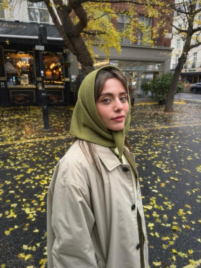 Девушка в зелёной косынке, завязанной как шарф, на фоне опавших жёлтых листьев; естественный уличный кадр с осенне-зимней атмосферой.