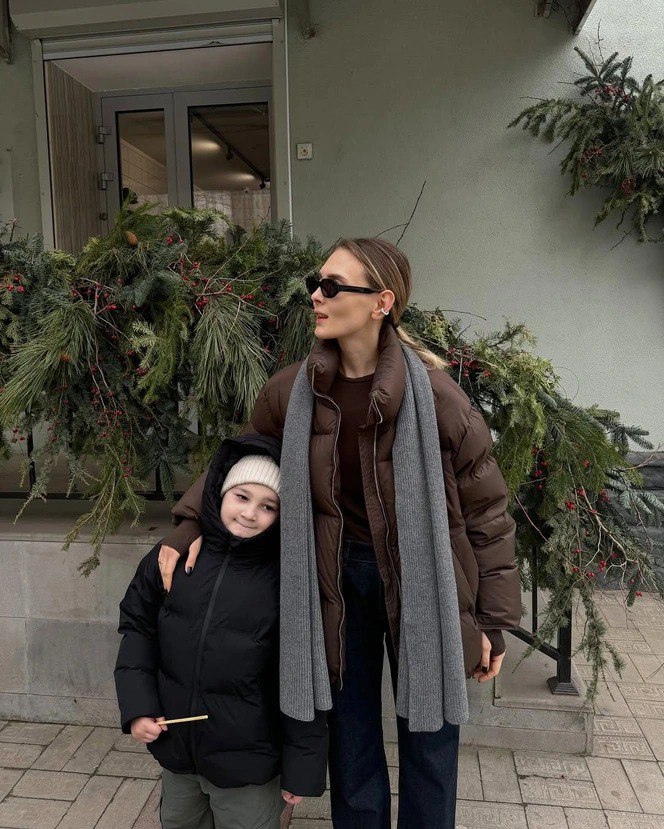 Мама с ребёнком в тёплых пуховиках: длинный тонкий шарф-снуд серого цвета, семейный уличный образ в зимней городской обстановке.