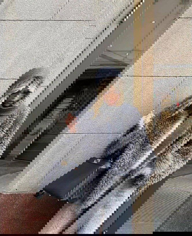 Пушистый серый шарф-шаль в сочетании с меховым пальто и джинсами; кадр у входа в магазин подчёркивает текстуру и материальность аксессуара.