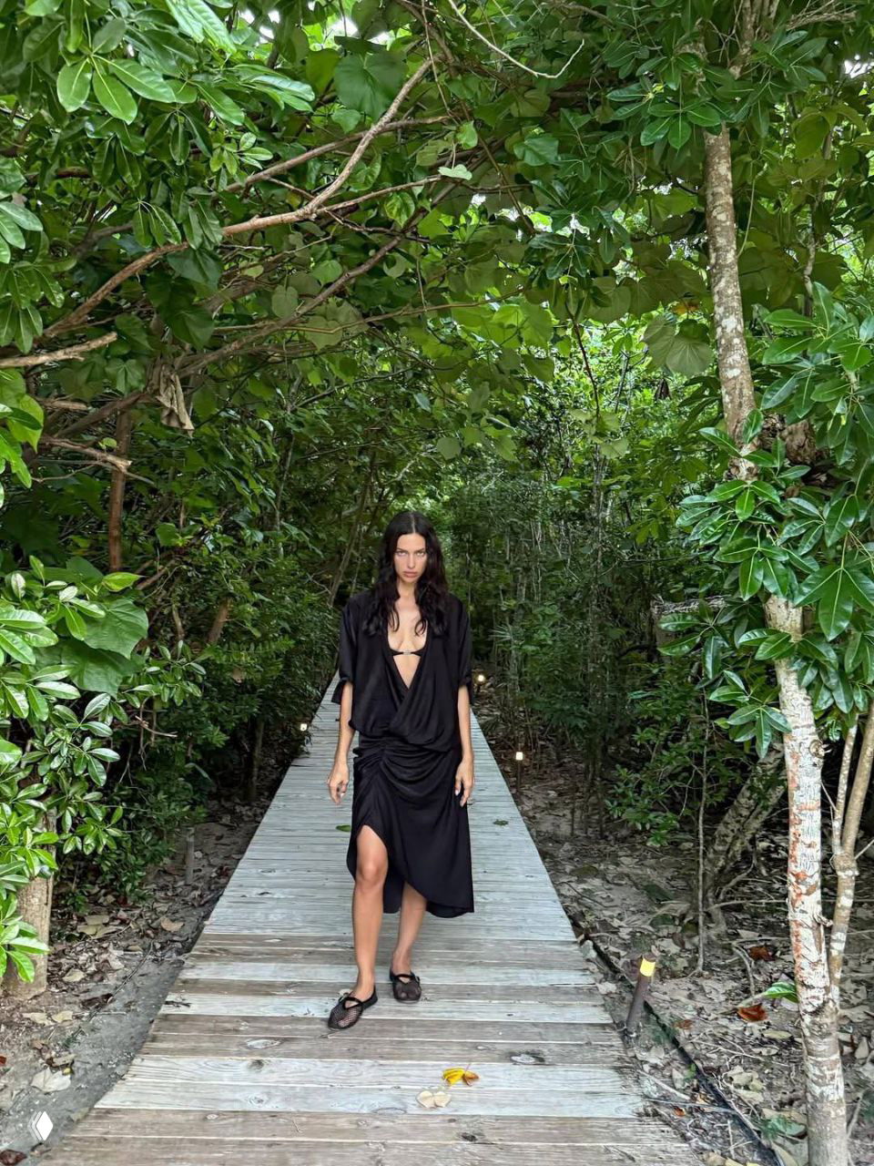Ирина Шейк проходит по настилу сквозь густую тропическую растительность, черное платье и расслабленная походка — атмосфера уединённого курорта.