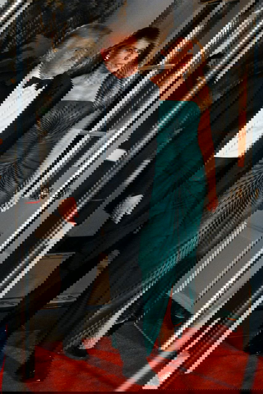 Женщина в изумрудном вечернем платье идёт по красной дорожке рядом с мужчиной в чёрном смокинге — момент прибытия на официальный приём в резиденции.