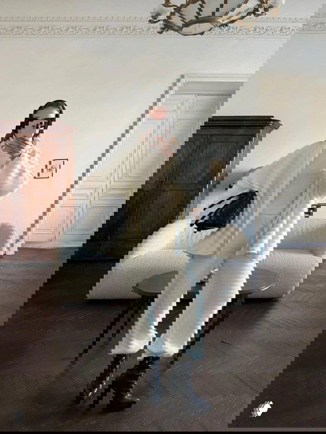 Женщина в тотал‑белом образе: объёмные вязаные белые брюки, свободный кардиган и высокие чёрные сапоги в элегантной гостиной с диваном и мебелью.