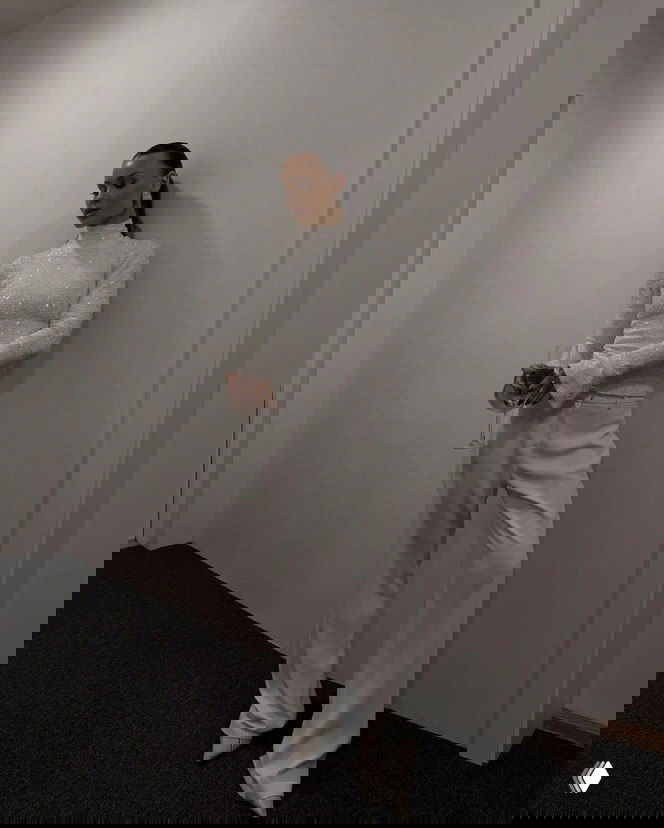 Модель в белых прямых брюках и блестящем светлом топе держит бокал, строгая поза на нейтральном фоне — идея праздничного образа с белыми брюками.