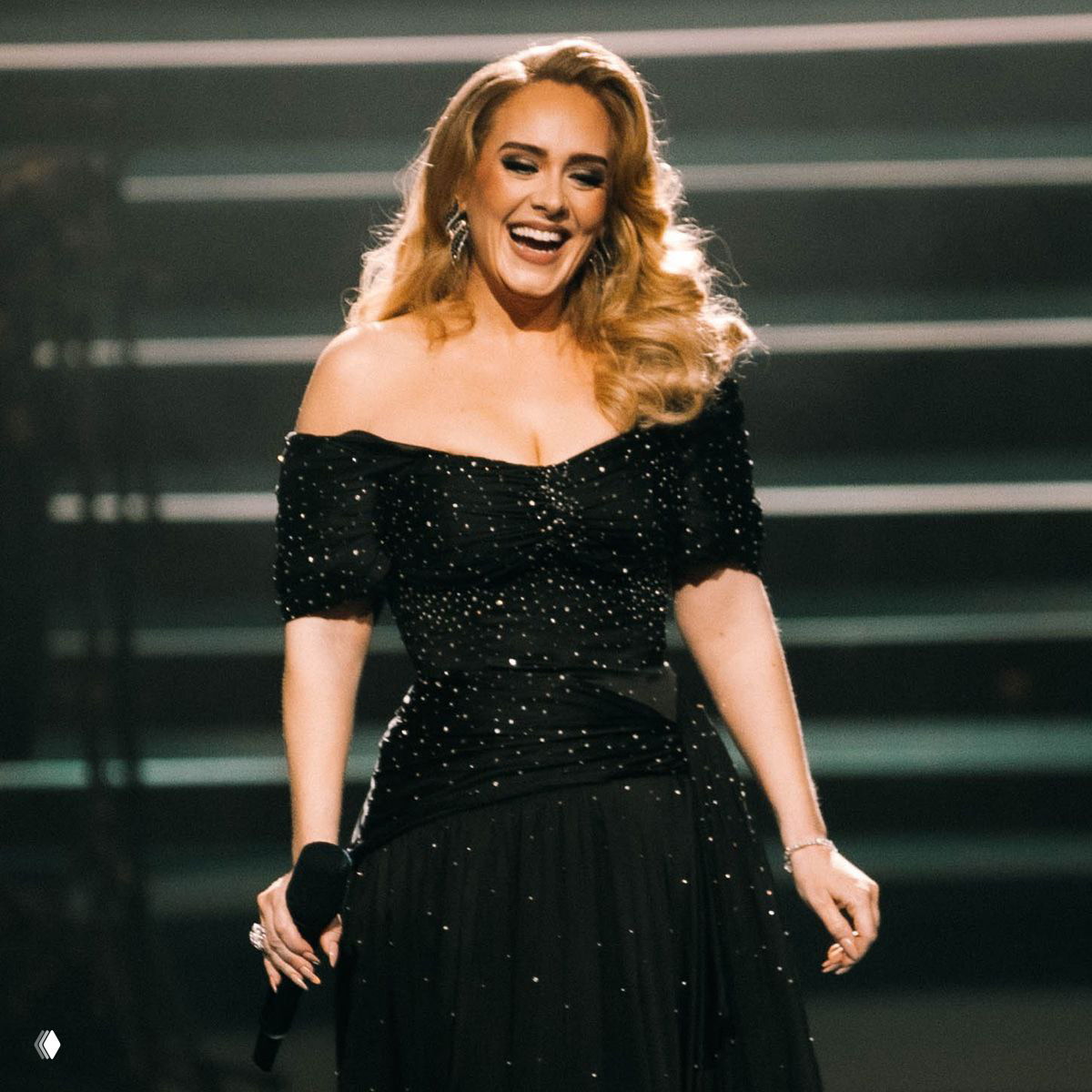 Певица на сцене в чёрном платье с микрофоном, улыбается; крупный план, театральное освещение и ступенчатый фон