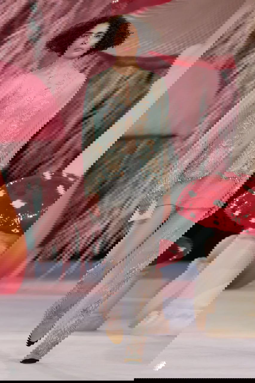 Модель в тонком полупрозрачном жакете и вышитых брюках шагает по подиуму Chanel на фоне пастельного лесного оформления показа.