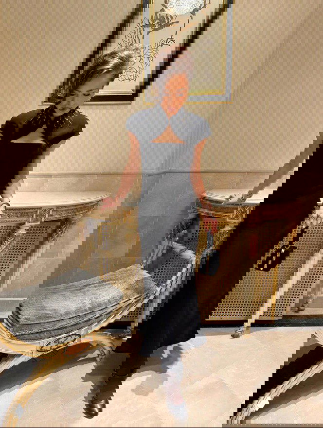 Женщина в длинном чёрном вечернем платье у изысканной консоли и стула в интерьере, подчёркнут элегантный образ и аксессуары