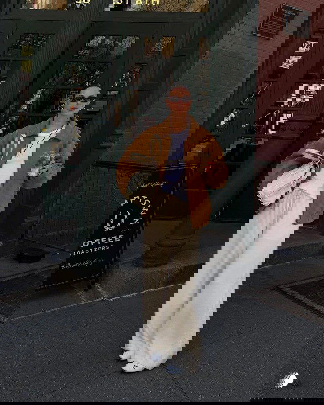 Уличный образ: широкие кремово‑белые брюки, коричневая куртка‑бомбер, белая сумка и кроссовки; девушка идёт по городскому тротуару с напитком в руках.