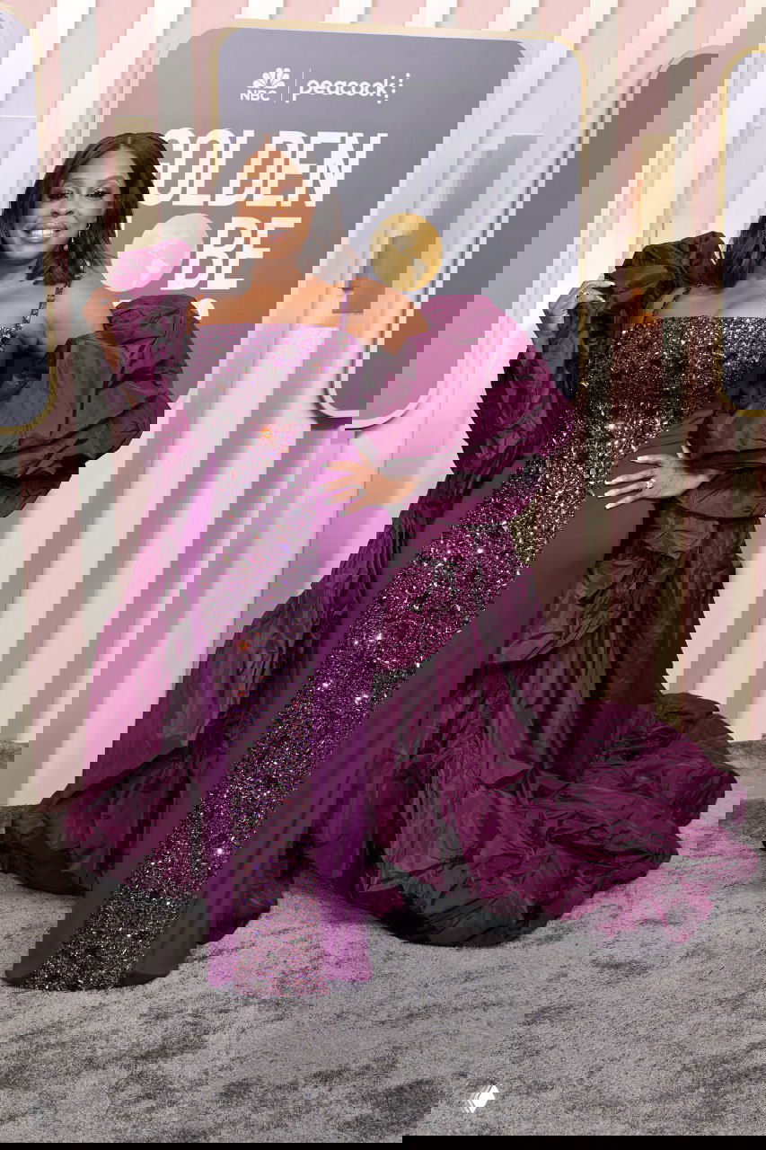 Полнофигурная женщина в роскошном глубокофиолетовом платье с пышной драпировкой и расшитым лифом, позирует на фотозоне с логотипом премии.