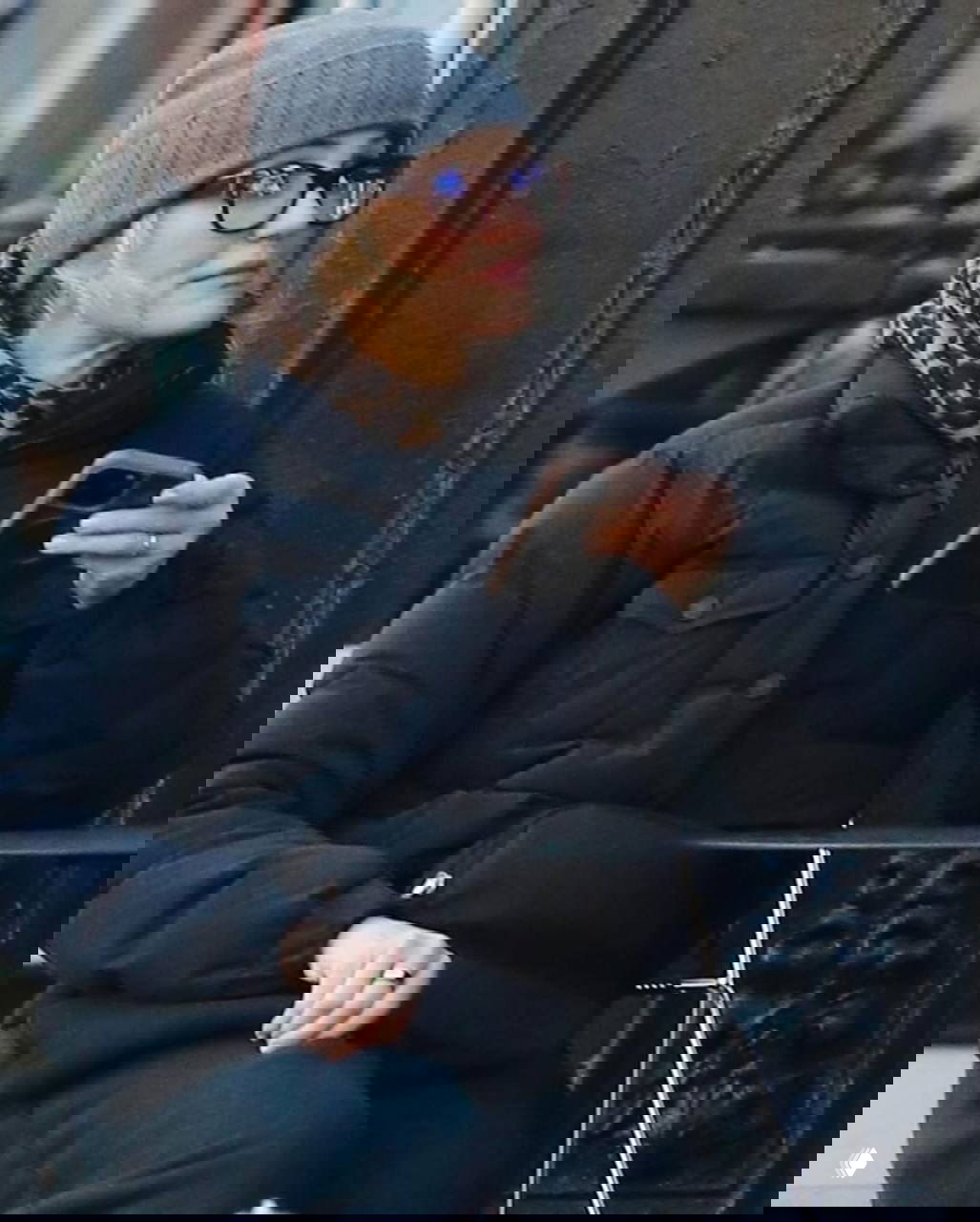 Женщина в серой шапке и тёплом пуховике с леопардовым шарфом сидит за уличным столиком и держит смартфон, повседневный зимний кадр.