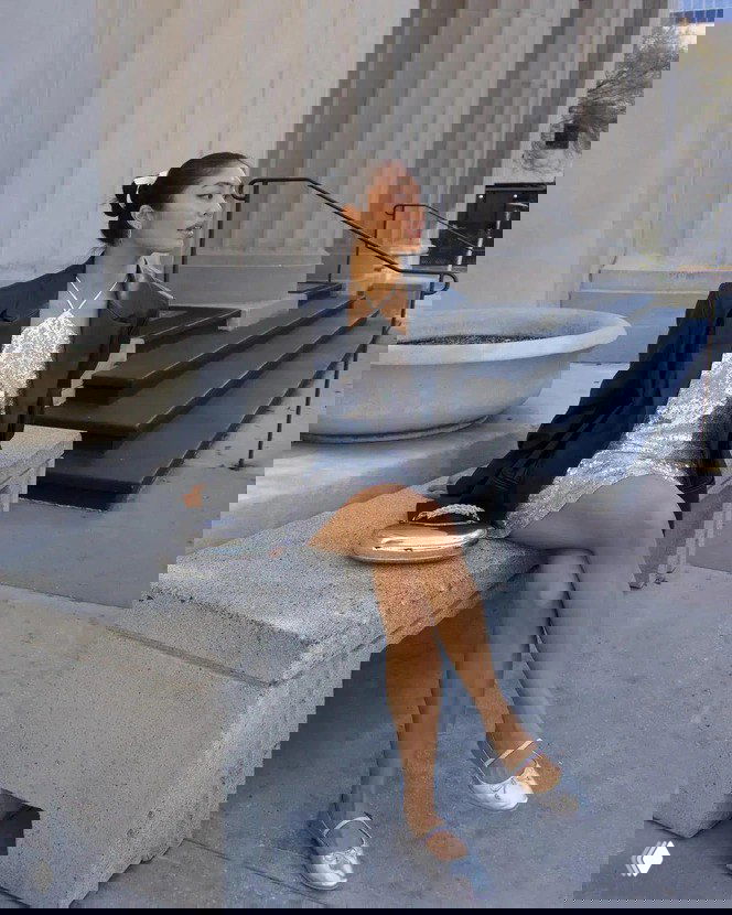 Девушка в серебристом блестящем мини-платье и чёрном пиджаке сидит на бетонной скамье у монументального здания, образ с балетками в уличном контексте