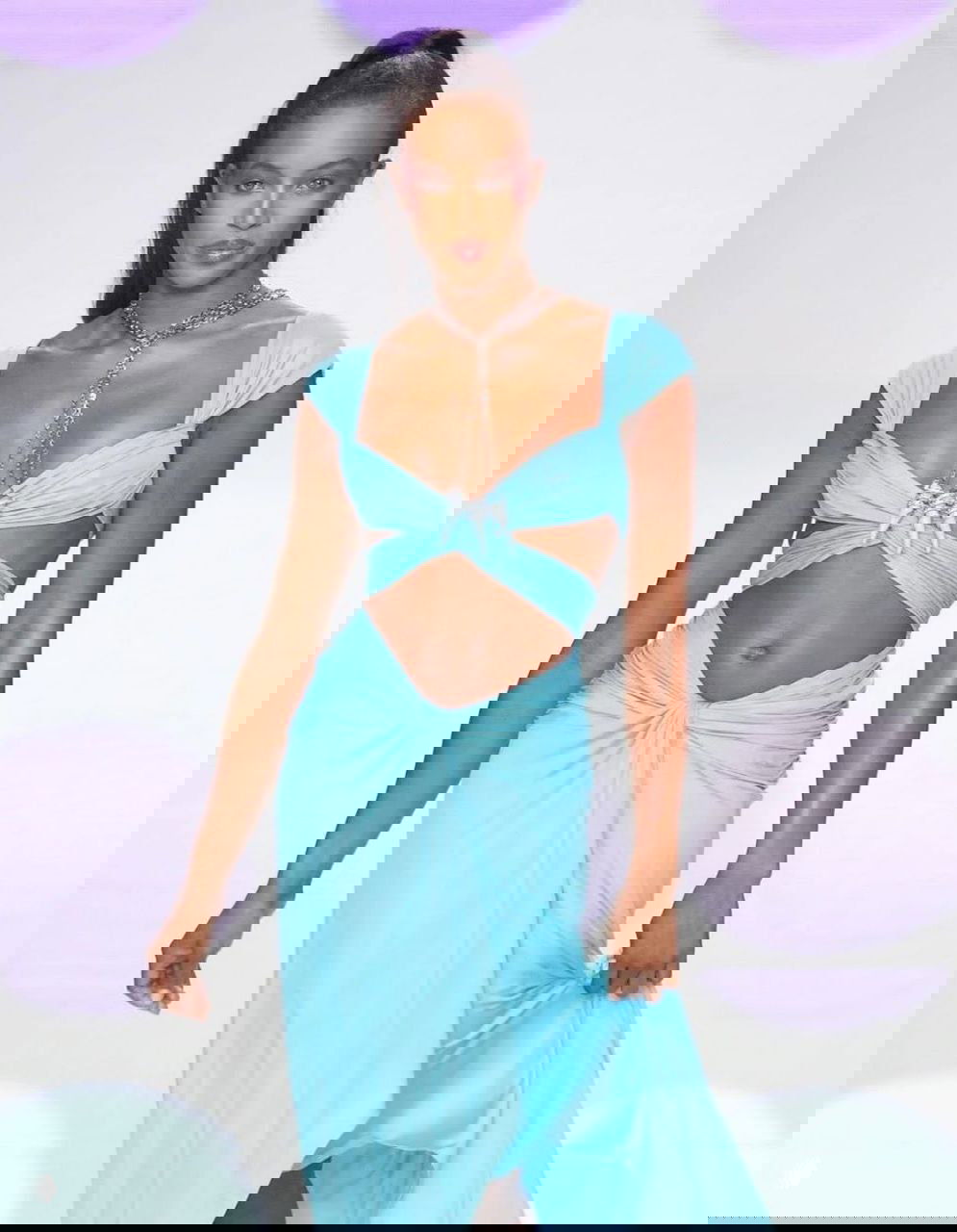 Модель в светло-бирюзовом разрезном платье идёт по взлётной дорожке, образ подчёркнут длинной цепочкой и макияжем.