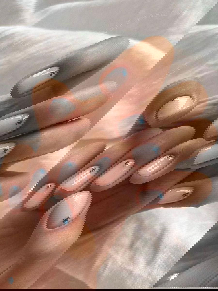 Пара рук с нежно-белым глянцевым маникюром и маленькими синими цветочками у кутикулы, композиция выложена на белом текстиле.