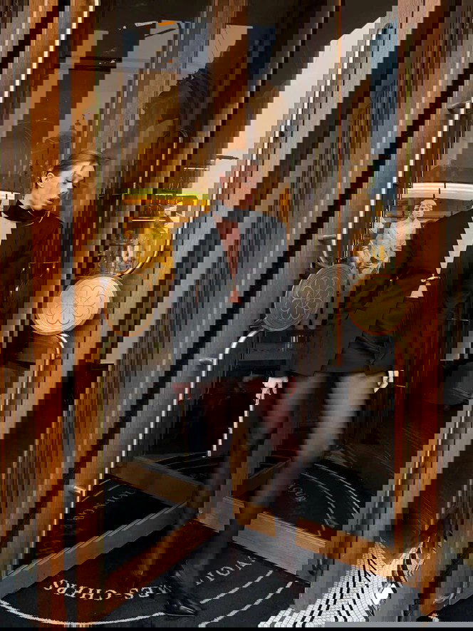 Женщина в чёрном жакете и мини-юбке выходит через вращающиеся двери отеля — образ с акцентом на силуэт и прозрачные чулки