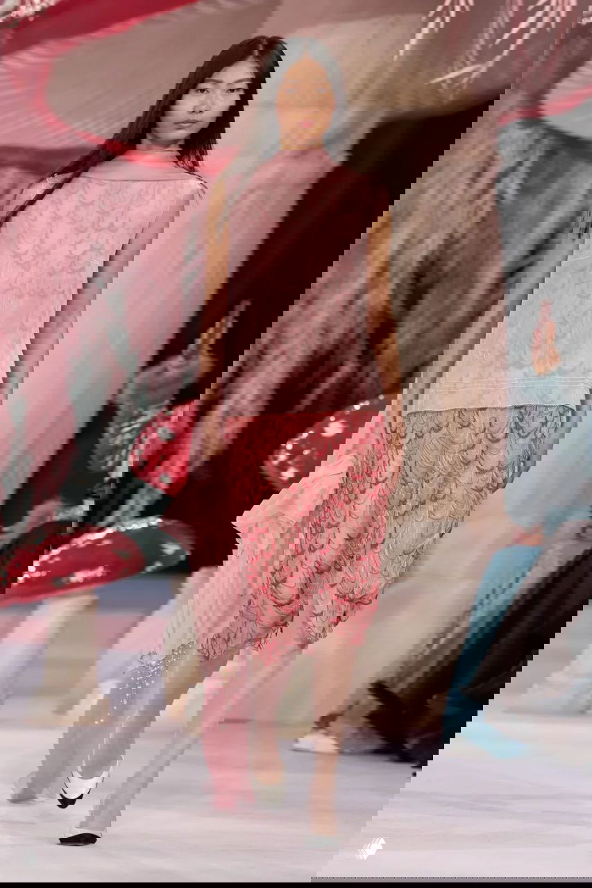 Модель в розово-персиковом комплекте с узором перьев идёт по подиуму Chanel, перед крупными красными грибами и пастельными декорациями.