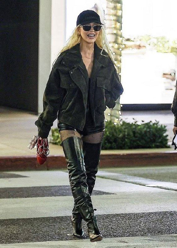 Женщина в чёрной короткой куртке, кожаных шортах и высоких ботфортах пересекает дорогу вечером, в кадре видны уличные огни и праздничная иллюминация.