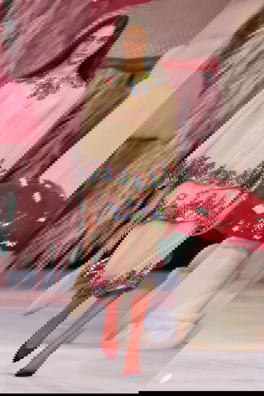 Модель в светло-жёлтом полупрозрачном костюме с цветочной отделкой шагает по подиуму Chanel на фоне сказочных грибов и розовых растений.