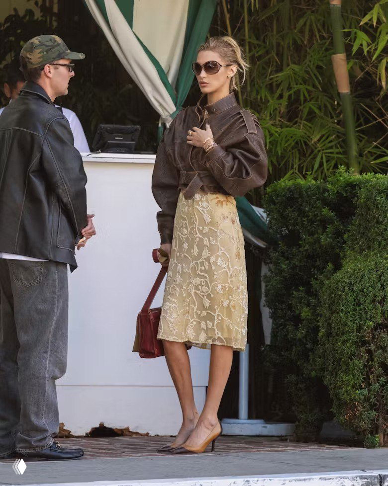 Белла Хадид у входа в кафе: тонкое светлое вышитое платье, объёмная коричневая кожаная куртка, солнцезащитные очки и красная сумка в руках.