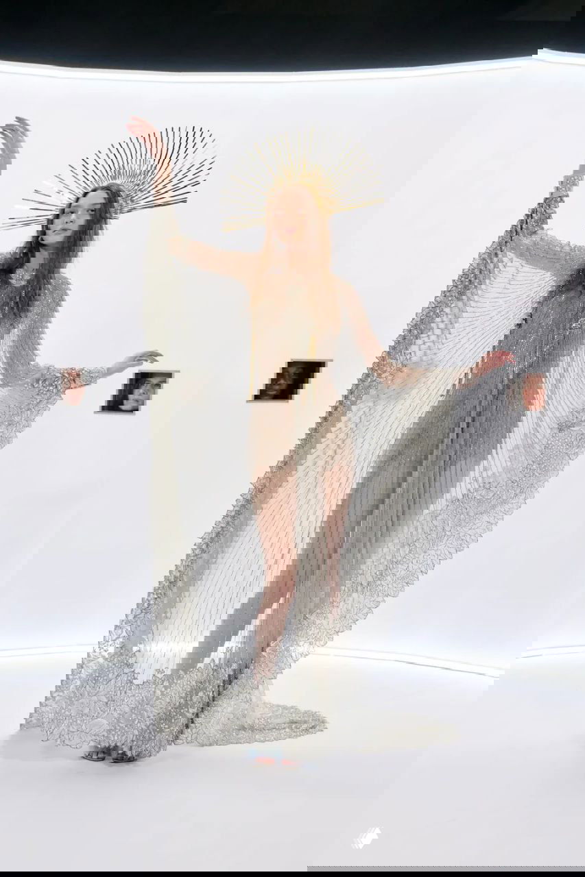 Модель в прозрачном золотистом платье и короне-ореоле, развёрнутая на белом подиуме с мини-портретами в стенах.