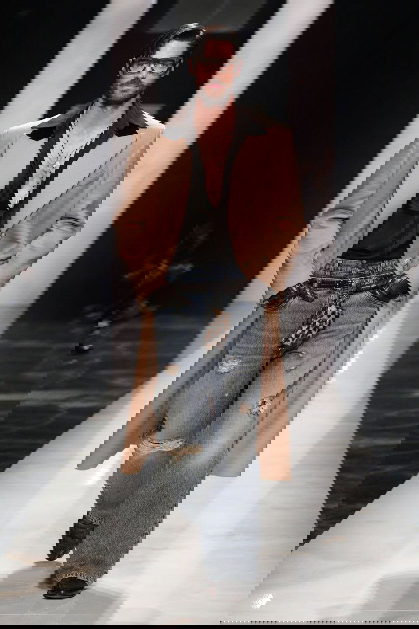 Мужчина в длинном бежевом пальто и рваных джинсах идет по подиуму Gucci — сочетание классики и небрежности в мужской моде осенью.