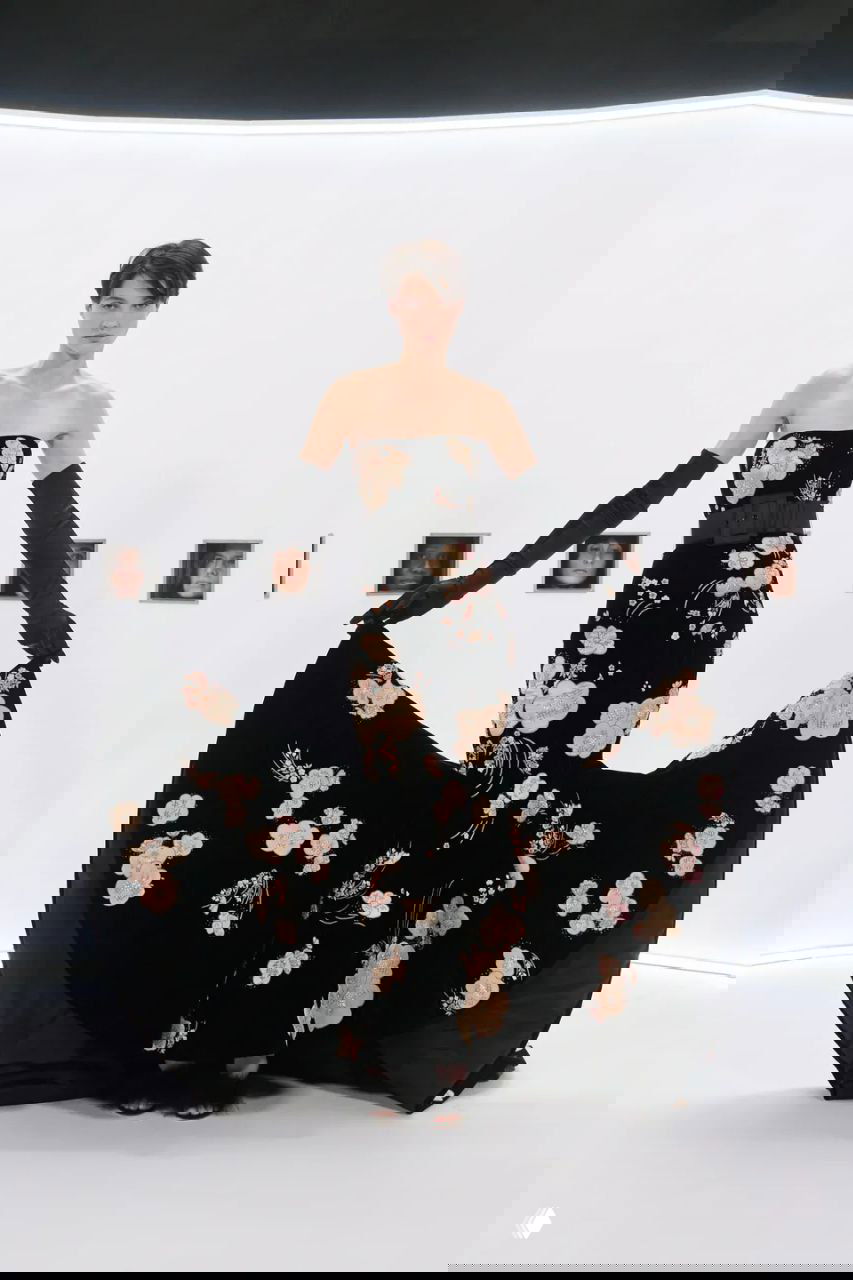 Модель в обтягивающем чёрном платье с цветочной вышивкой и широкой юбкой, позирующая в круглом белом пространстве показа.