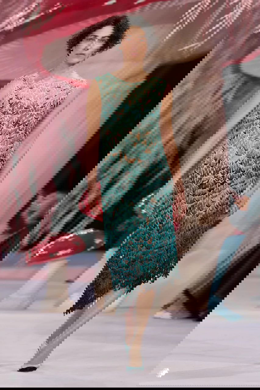 Модель в ажурном платье бирюзовых оттенков идёт по подиуму Chanel на фоне сценографии с большими грибами и розовыми элементами «лесной» темы.