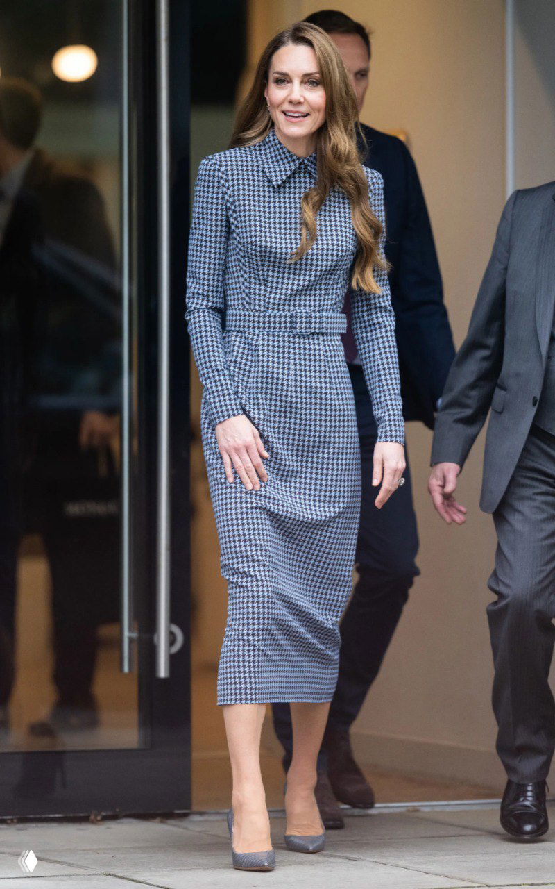 Женщина в платье в ломаную клетку и серых туфлях выходит из здания, улыбается и демонстрирует строгий, но элегантный образ на фоне улицы