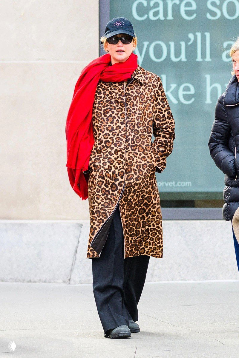 Женщина на городской улице в длинном леопардовом пальто и тёмных брюках, с ярким красным шарфом; уличный кадр во время прогулки по городу.