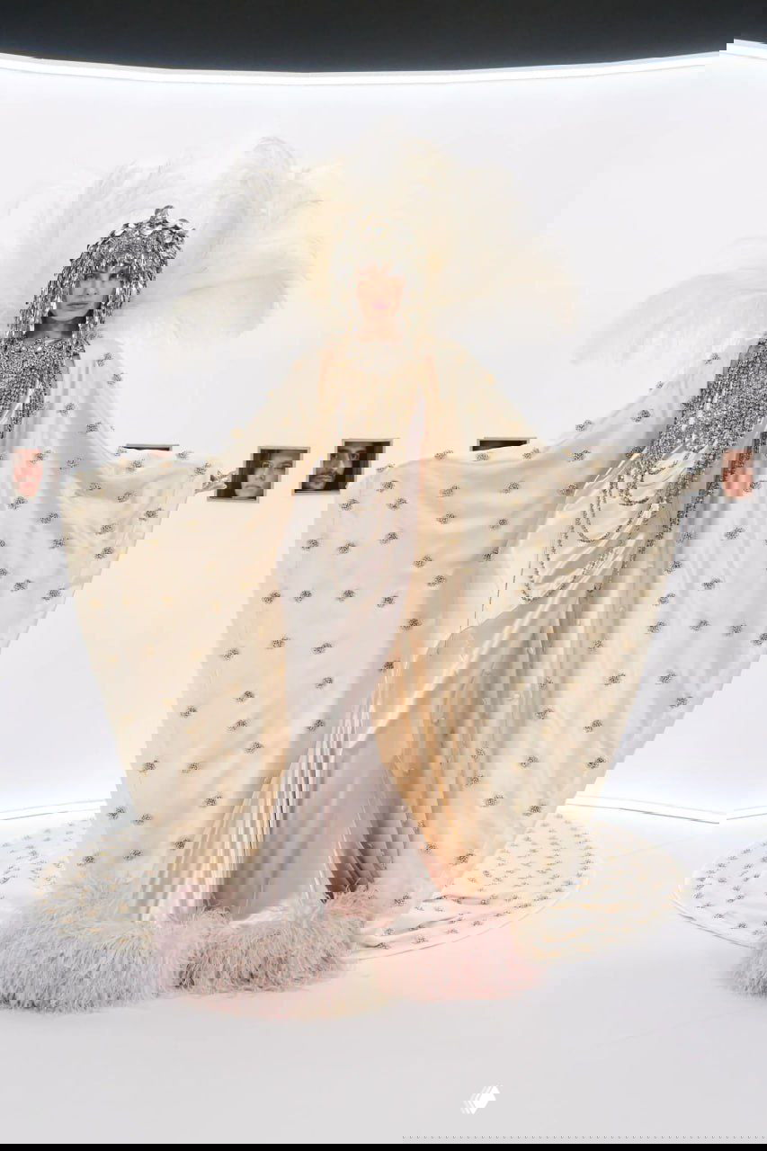 Авангардный образ: светло-бирюзовая юбка-складки с чёрными рюшами и крупным перьевым аксессуаром на голове в белом пространстве показа.