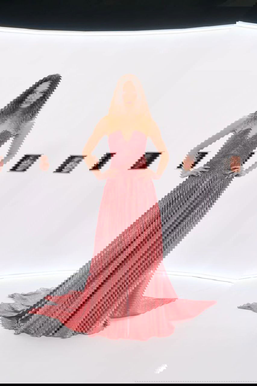 Модель в светло-бежественном платье с длинной мантией и обильными украшениями, стоящая в центре круглой белой сцены с мелкими портретами.